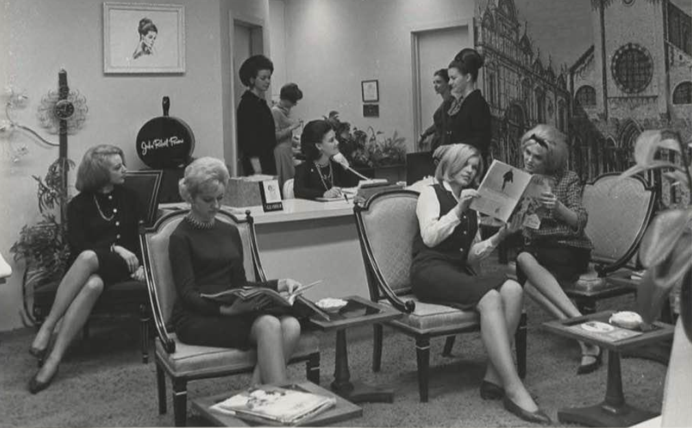 Black and white photo of young women standing, sitting, and reading in an office.